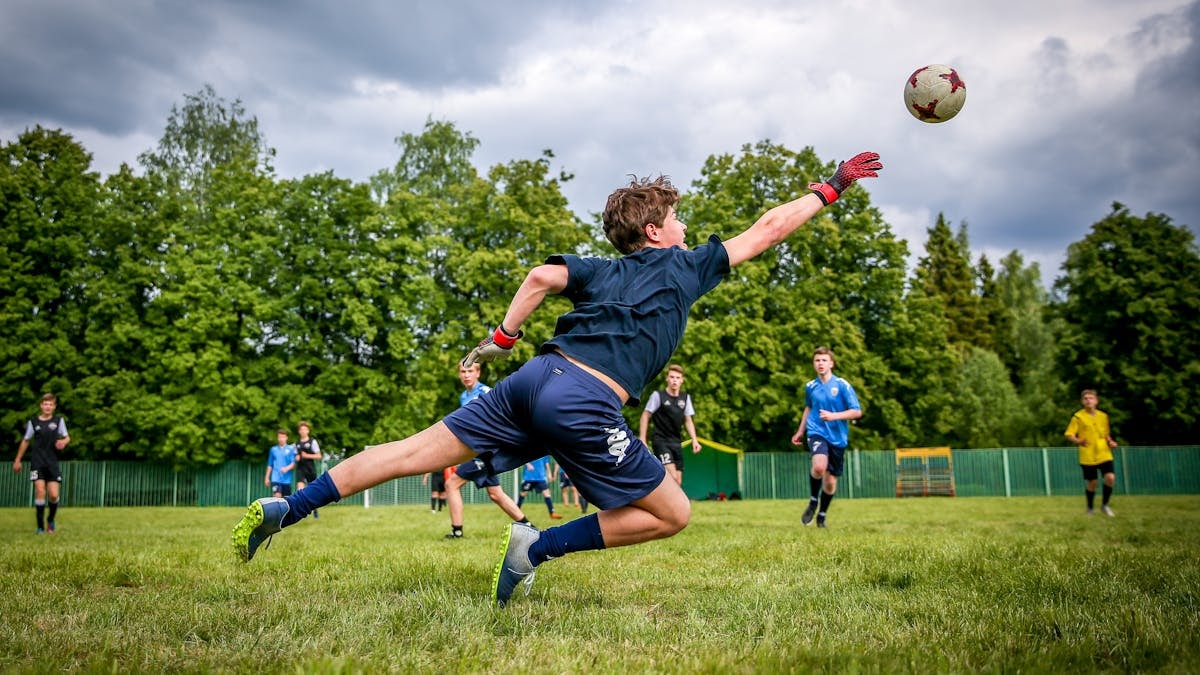 A young goalkeeper dives to catch a soccer ball during a lively outdoor game on a green field.